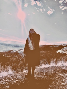 A woman in a large cardigan stands on a mountain at sunset. 
