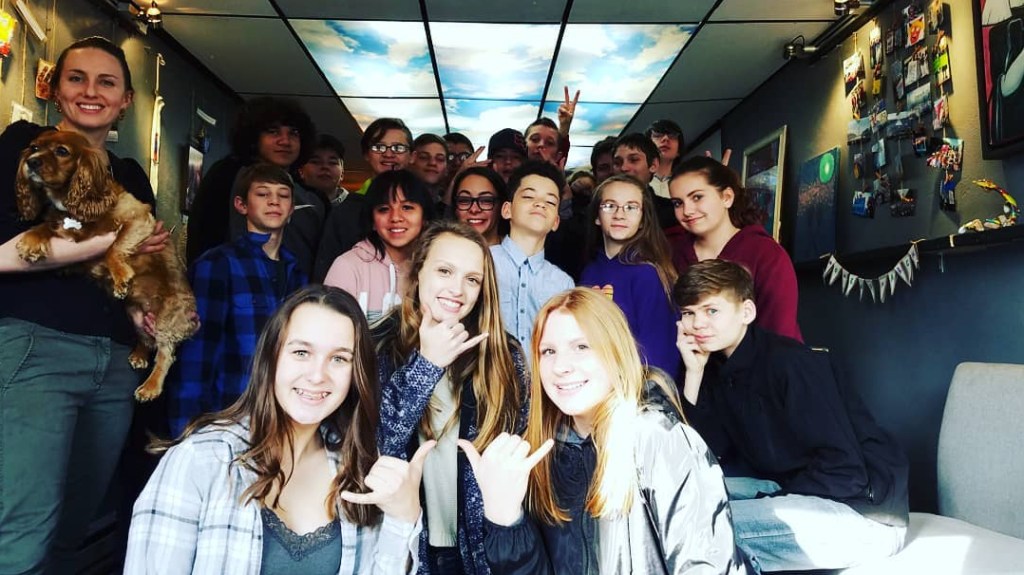a class of eighth graders stand in a gallery with grey walls and art hanging on the walls. They all smile towards the camera, and an adult woman holds a dog and is smiling too.