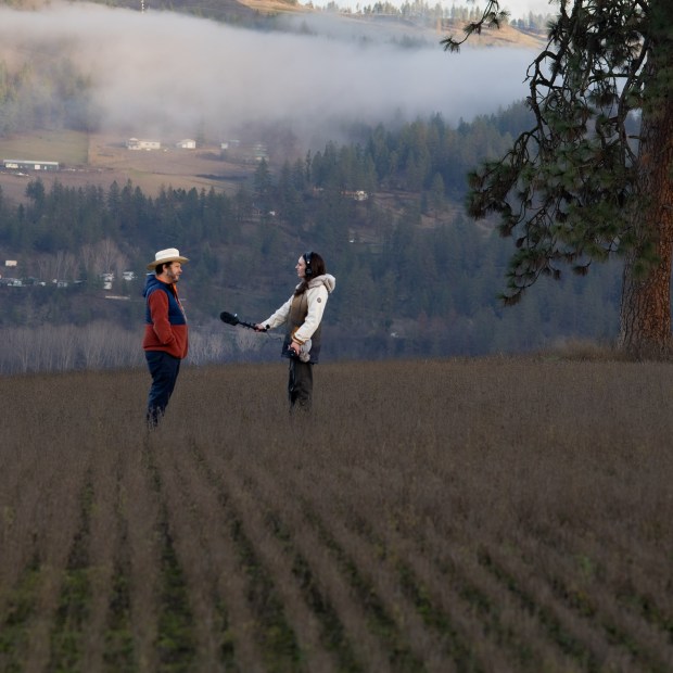 A woman in a jacket with a microphone interviews a man in an orange jacket and cowboy hat across from her against a mountain landscape while standing in a field.